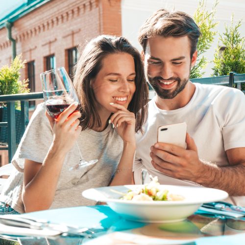 Smiling beautiful woman and her handsome boyfriend. Couple cheering with glasses of red wine at their date in restaurant. They drinking alcohol at veranda cafe in the street.Looking at phone screen