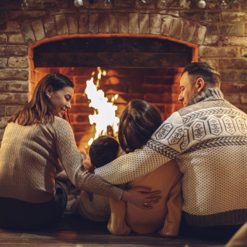 Happy young family parents with two small kids gathering around brick fireplace in cozy and warm country house during Christmas holidays in winter. Father, mother ad children sitting by fire at home