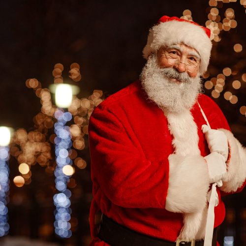Horizontal medium portrait of joyful Santa Claus standing outdoors in street decorated with garland lights smiling at camera, copy space